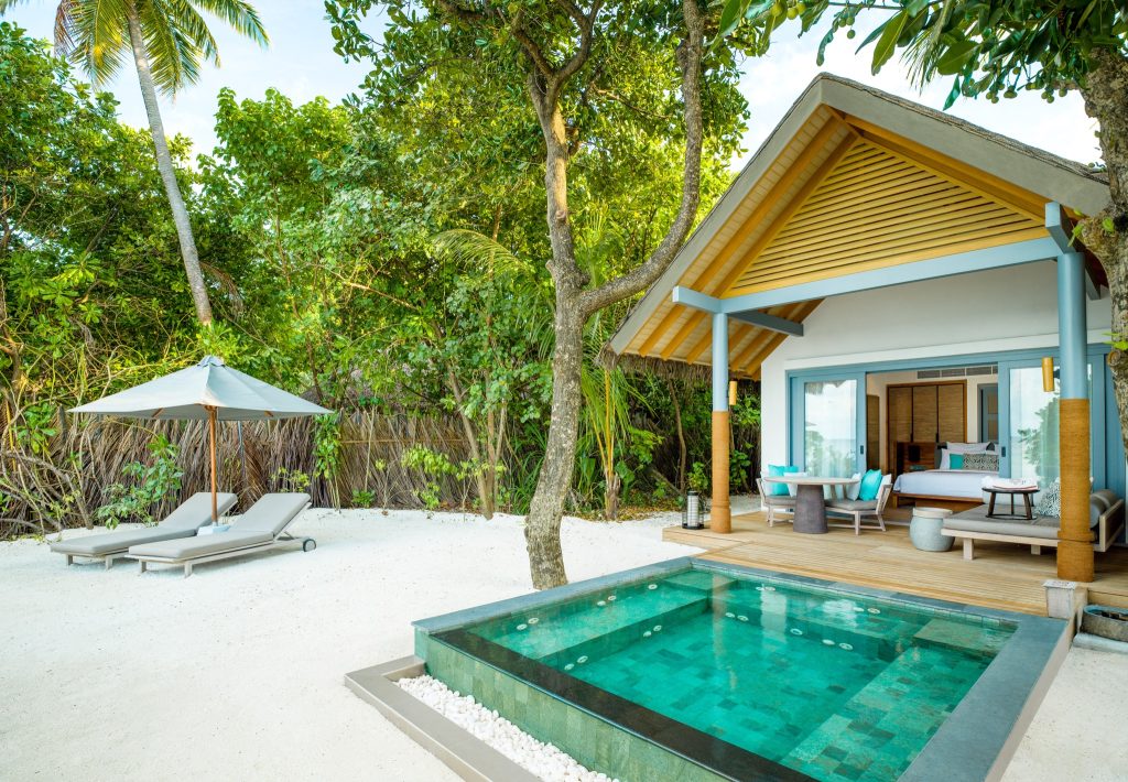 Tropische Villa am Strand mit Holzterrasse, kleinem Pool, blauen Sitzkissen und grauen Liegen auf Sand