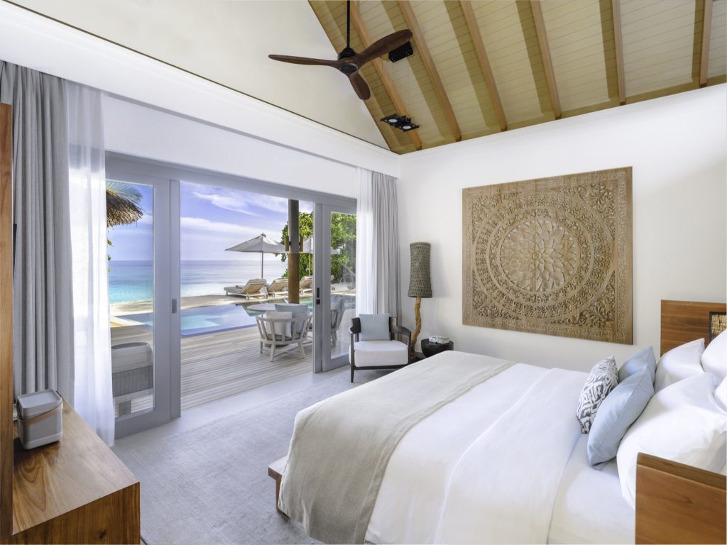 Modernes Hotelzimmer mit großem weißen Bett, blauen und beigefarbenen Kissen, Holz-Wandrelief und Meerblick durch Glasfront