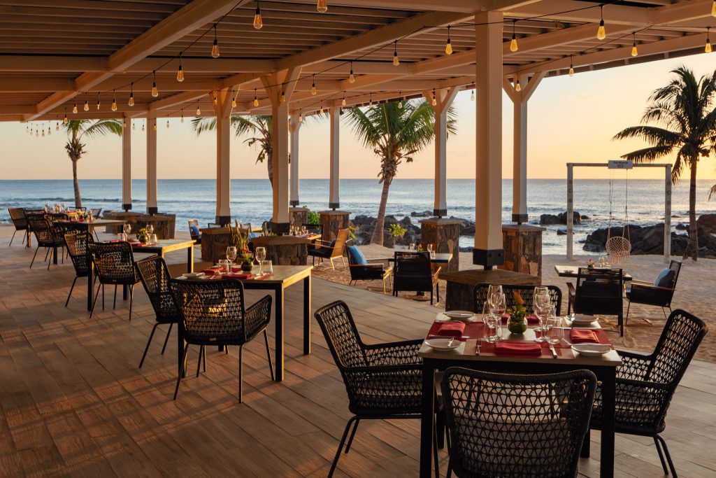 Offene Terrasse der Bar Mystique mit gedeckten Tischen, Stühlen, Palmen und Blick auf Meer und Sonnenuntergang