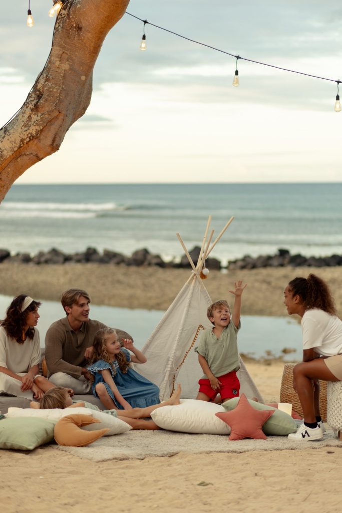 Fünf Personen sitzen auf Kissen und Teppich unter hellem Stoffzelt am Strand mit Baum und Meer im Hintergrund