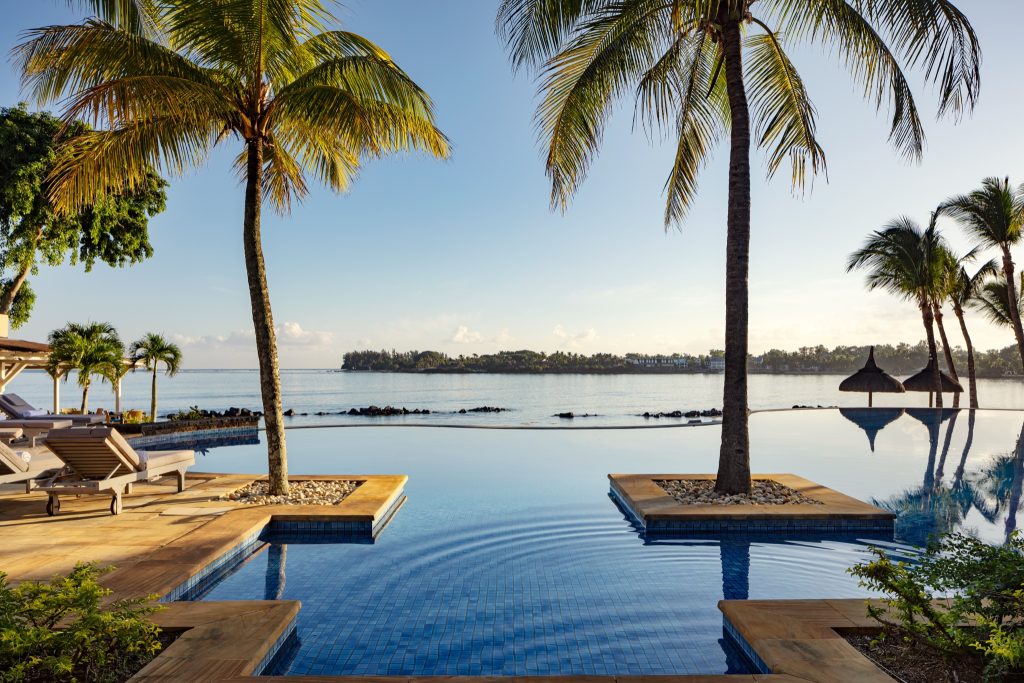 Infinity-Pool mit hellen Holzdielen und zwei Palmen in quadratischen Kiesbereichen am Meer bei klarem Wetter