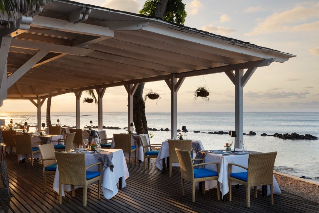 Überdachte Terrasse des Beach Grill Restaurants mit Holzboden, blauen Stühlen und Blick aufs Meer bei Sonnenuntergang
