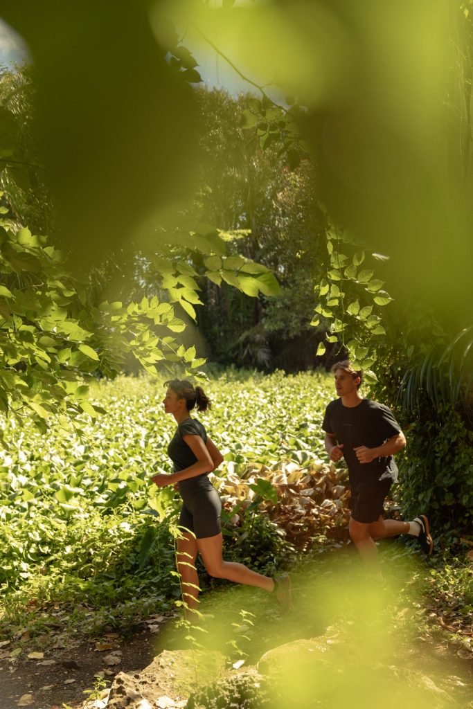 Zwei Personen laufen auf einer Laufstrecke durch einen grünen Wald mit Sonnenlicht und Blättern im Vordergrund