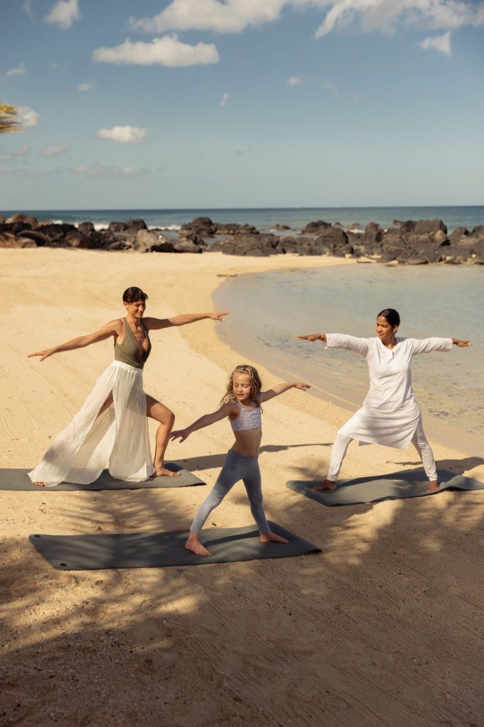 Drei Personen in weiß-beiger Kleidung machen Yoga am Sandstrand mit blauem Ozean und dunklen Felsen im Hintergrund
