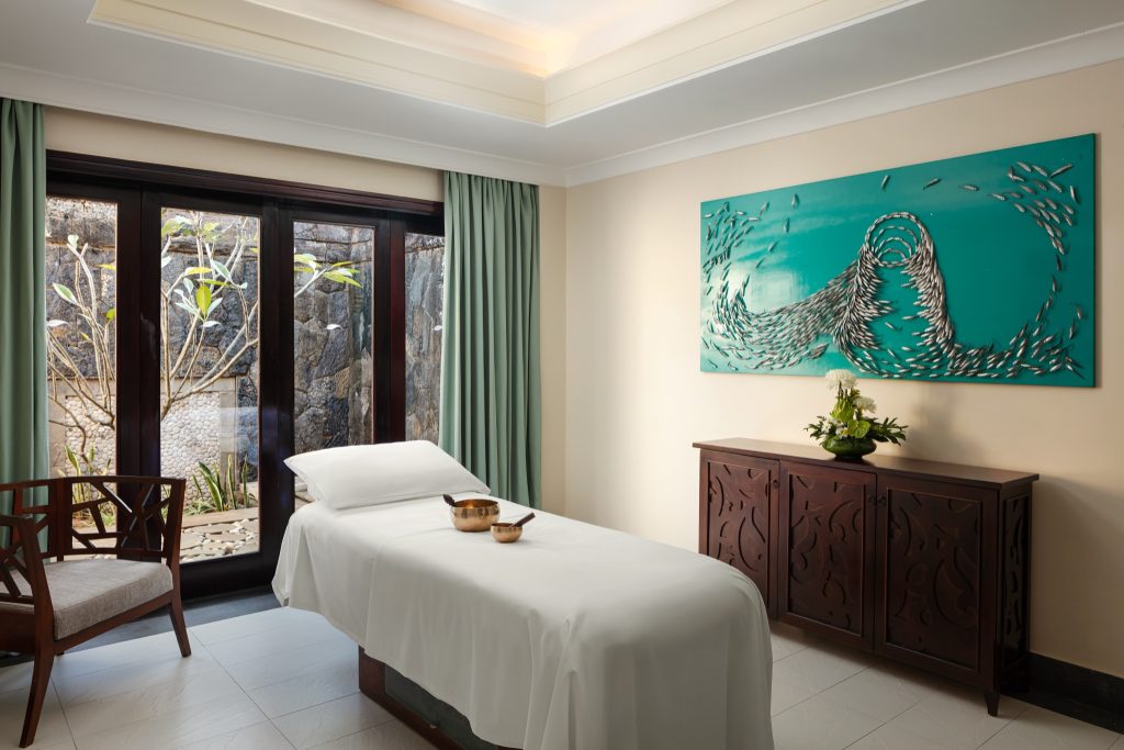 Helles Spa-Zimmer mit weißer Massageliege, goldfarbenen Schalen, dunkler Kommode und großen Fenstern