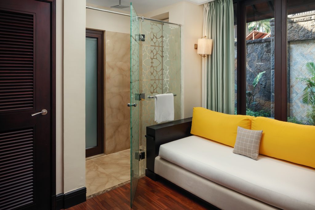 Banyan Suite mit beigem Sofa, gelben Kissen, bodentiefen Fenstern und Dusche mit Glastür