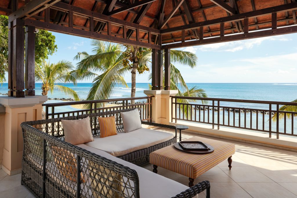 Offene Hotelterrasse mit dunklen Holzbalken, Rattansofa mit beigen Kissen und Meerblick im Hintergrund
