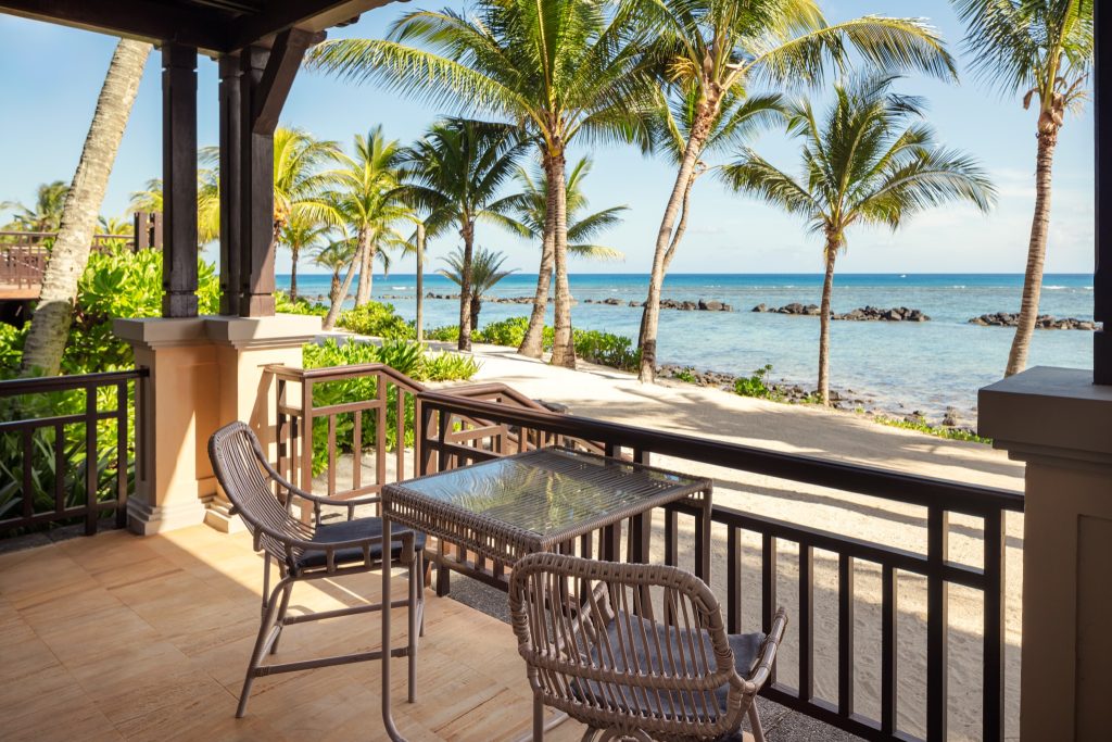 Terrasse mit Glastisch und zwei Stühlen aus hellem Flechtmaterial, Blick auf Strand und blaues Meer