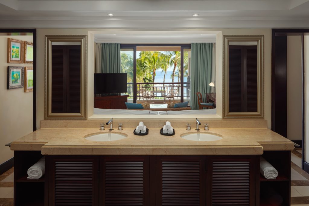 Badezimmer mit Doppelwaschbecken aus hellem Marmor, Spiegelblick ins Schlafzimmer mit Balkon und Meerblick