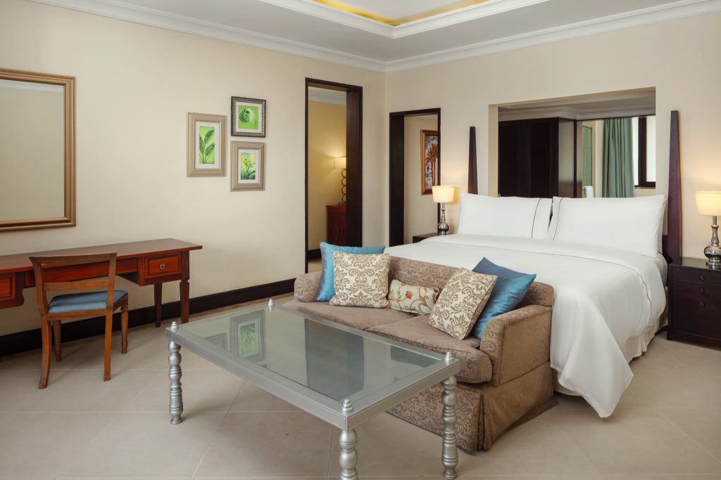 Zimmer der Turtle Bay Junior Suite mit großem Bett, gepolstertem Sessel, Glastisch und Holztisch in warmem Licht