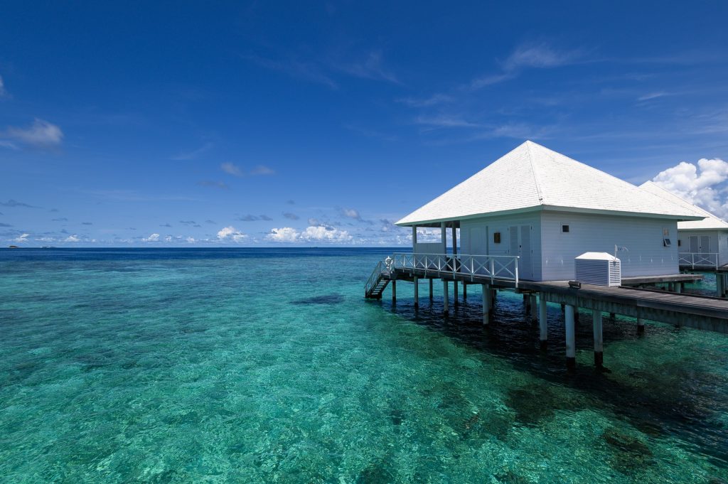 Überwasserbungalow mit weißem Dach auf Stelzen über türkisfarbenem Wasser, Holzterrasse und Treppe ins Meer