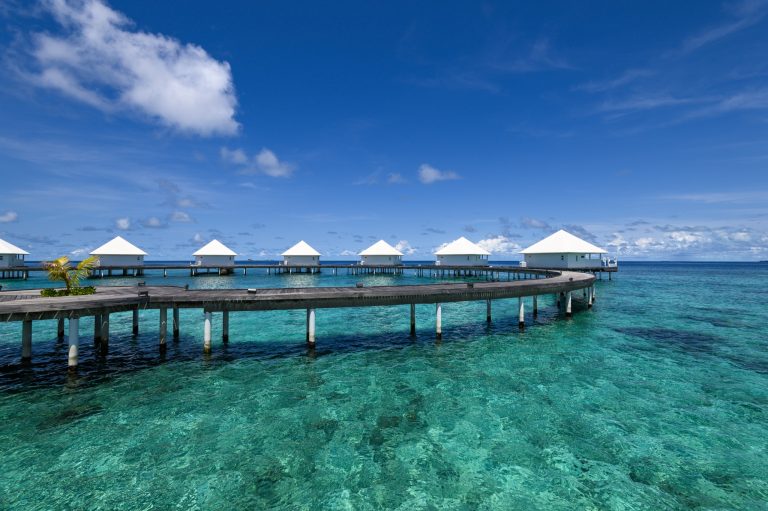 Reihe von Überwasserbungalows mit weißen Dächern auf Holzstegen über türkisfarbenem Meer bei klarem Himmel