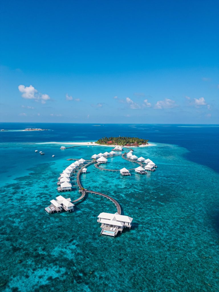Überwasser-Bungalows mit weißen Dächern auf Stelzen über türkisblauem Wasser, tropische Insel im Hintergrund