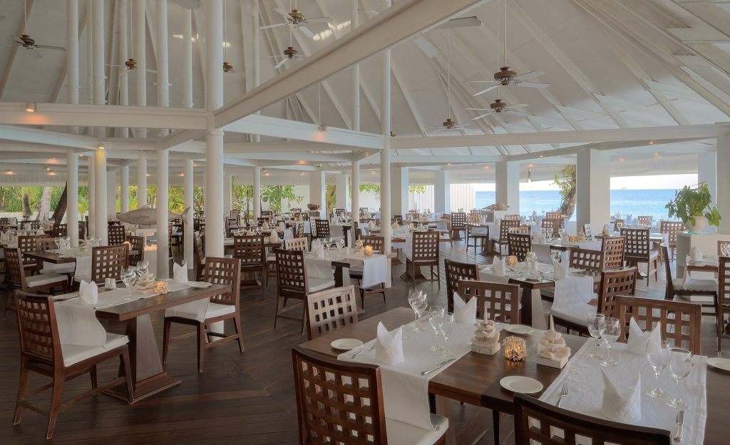Offenes Restaurant mit dunklen Holztischen, weißen Tischdecken, gitterartigen Holzstühlen und Blick auf das Meer