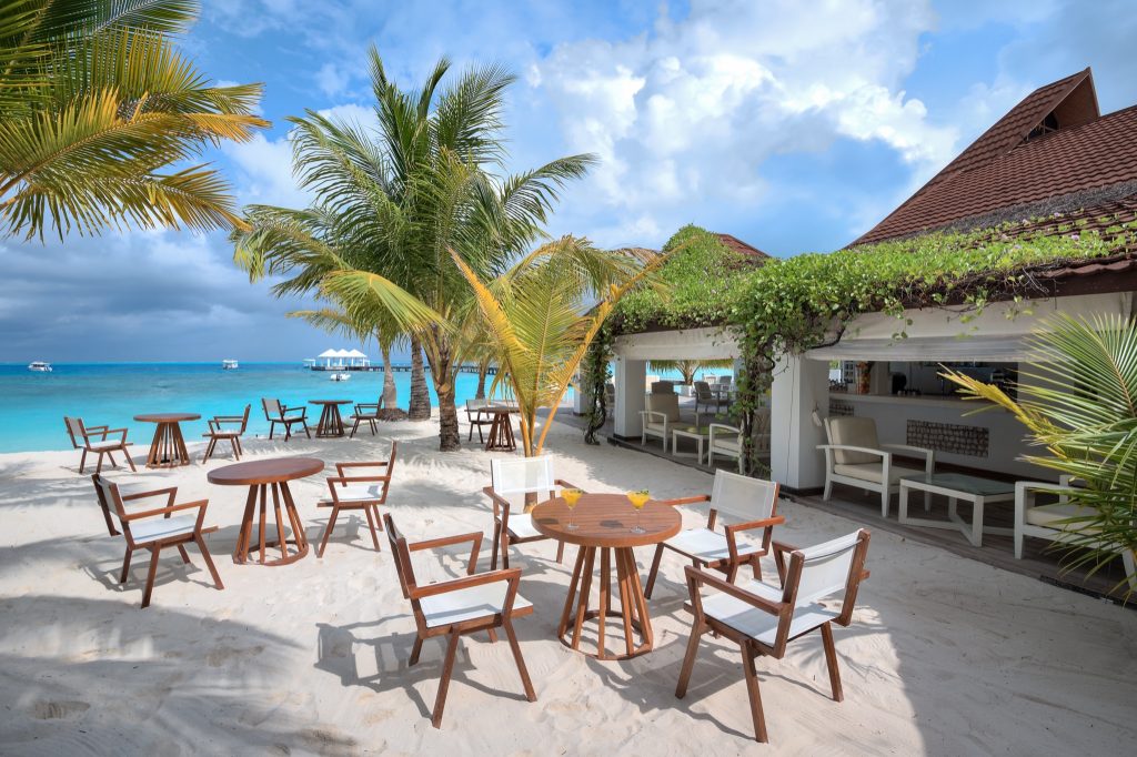 Strandrestaurant mit Holzstühlen und Holztischen auf weißem Sand, Palmen und türkisfarbenem Meer im Hintergrund