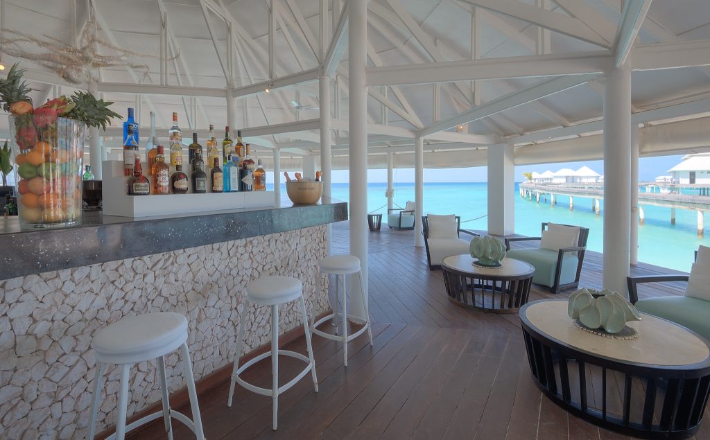 Offene Hotelbar mit dunkler Stein-Theke, weißen Barhockern und Blick auf türkisfarbenes Meer