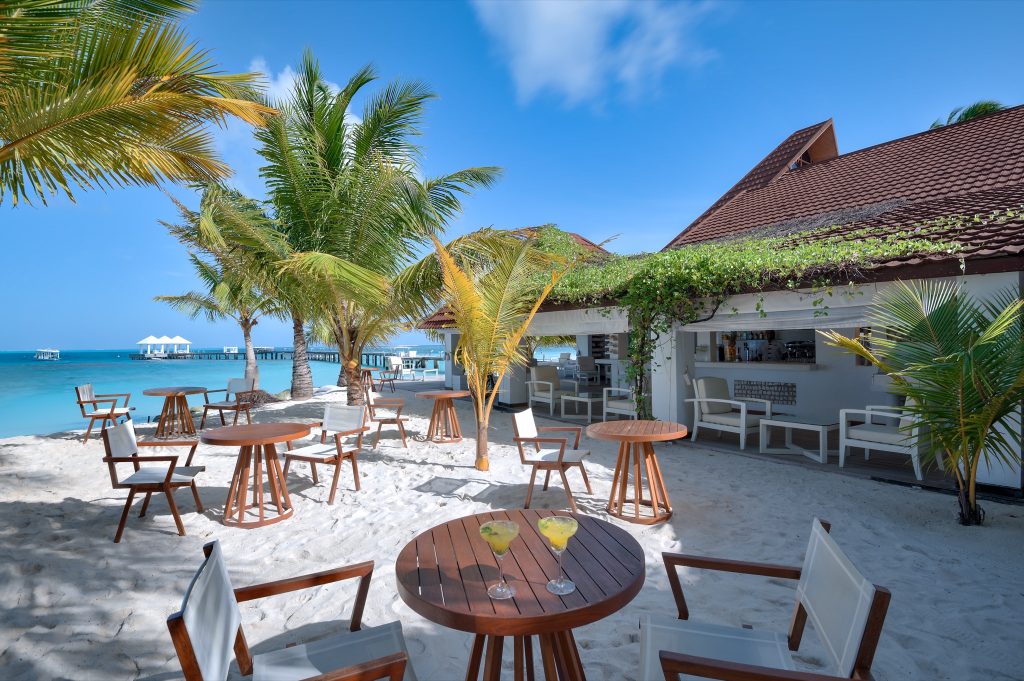Strandbereich mit runden Holztischen und weißen Stühlen vor offener Bar und tropischer Vegetation auf den