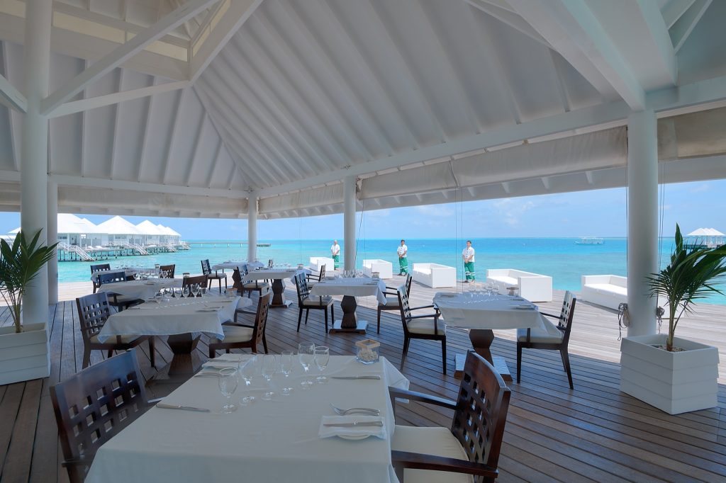 Außenrestaurant mit weiß gestrichenem Holzdach, Holztischen mit weißen Tischdecken und Blick auf das blaue Meer