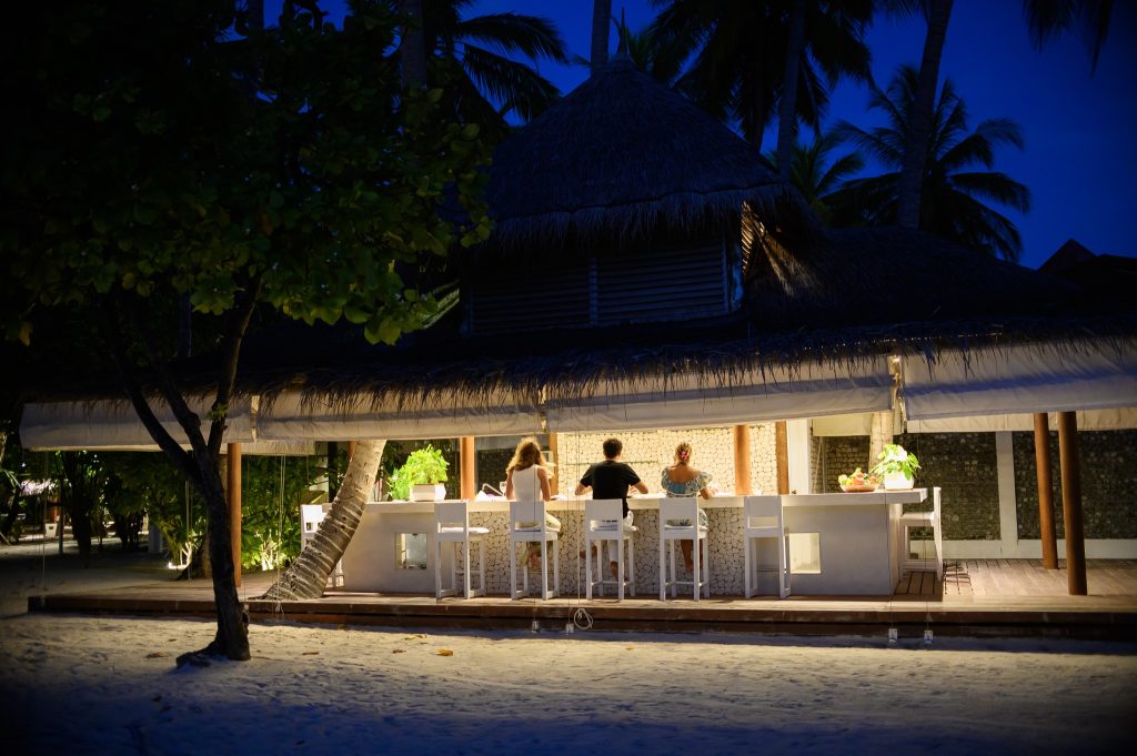 Strandbar mit Strohdach, drei Personen auf weißen Barhockern, beleuchtete Theke, Palmen im Hintergrund.