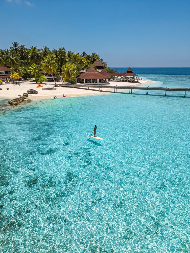 Resort mit spitz zulaufenden Dächern, Palmen, Steg ins türkisfarbene Meer und Stand-up-Paddler