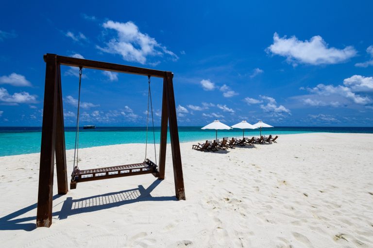 Weißer Sandstrand mit Holzschaukel und Liegestühlen unter weißen Sonnenschirmen am türkisfarbenen Meer
