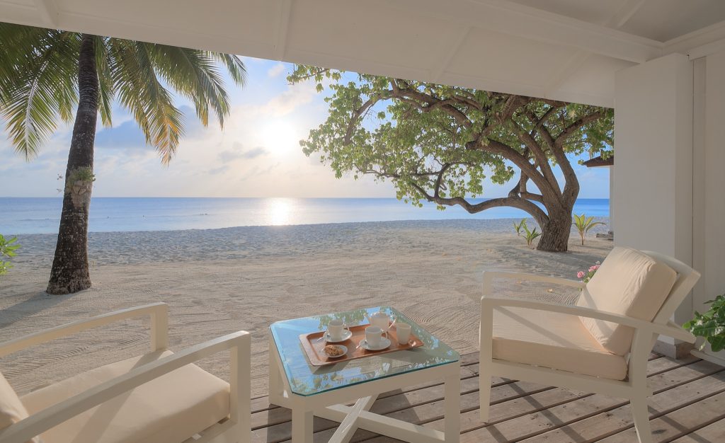 Terrasse mit weißen Stühlen, kleinem Glastisch und Tablett mit Tassen, Blick auf Strand, Meer und Palmen
