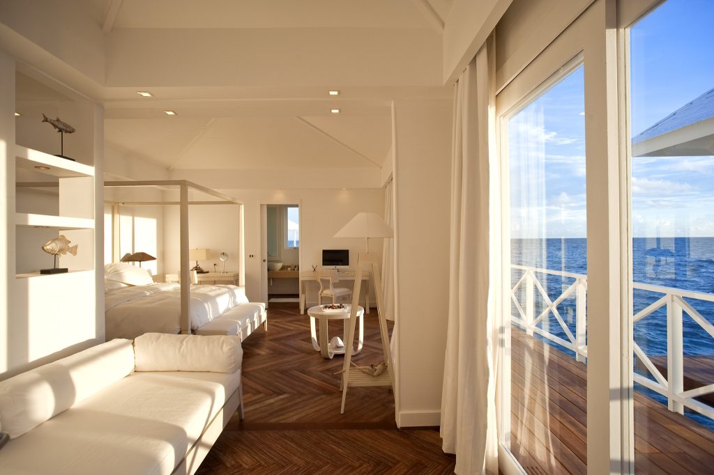 Helles Zimmer mit Himmelbett, Fischgrät-Holzboden und Meerblick durch großes Fenster im Diamonds Thudufushi Maldives Resort.