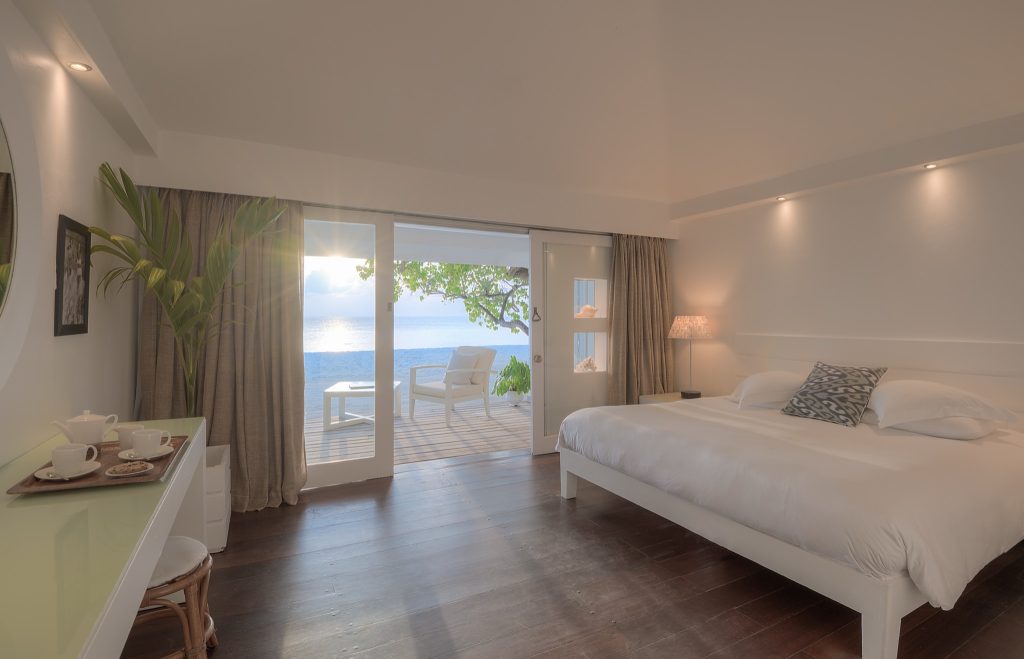 Helles Hotelzimmer mit großem Bett, weißen Bettlaken, minimalistischer Tisch mit Teekanne, Pflanze und Terrasse zum Ozean