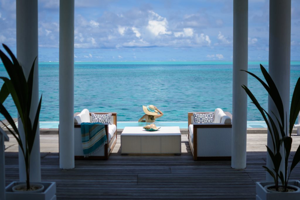 Terrasse mit zwei weißen Sofas, Holzelementen, weißem Tisch mit Muschel, Pflanzen und Blick auf türkisblaues Meer