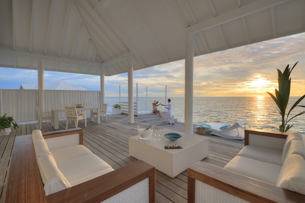 Offene Terrasse mit weißen Sofas, Rattan-Couchtisch, Holzboden und Meerblick bei Sonnenuntergang