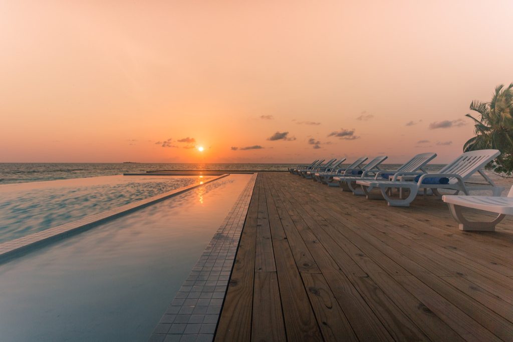 Infinity-Pool mit hölzernem Boden und weißen Sonnenliegen, Sonnenuntergang mit orangefarbenem Himmel auf den
