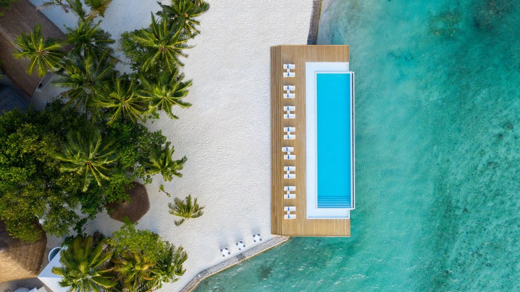 Luftaufnahme des Sandies Bathala Maldives Pools mit türkisblauem Wasser, Holzterrasse, weißen Liegestühlen und angrenzendem.