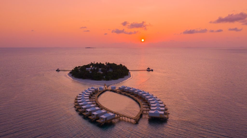Luftaufnahme von über Wasser gebauten Bungalows auf Stelzen mit Stegen und bewaldeter Insel im Sonnenuntergang