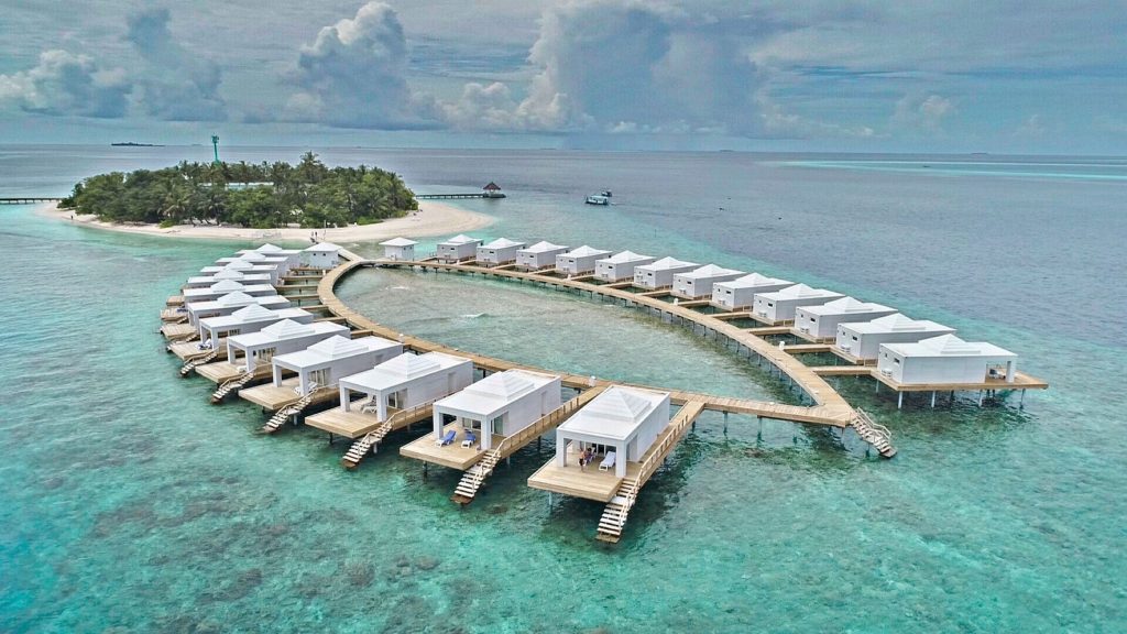 Weiße Überwasser-Bungalows auf Holzplattformen verbunden durch Stege vor bewucherter Insel und blaugrünem Wasser auf den.
