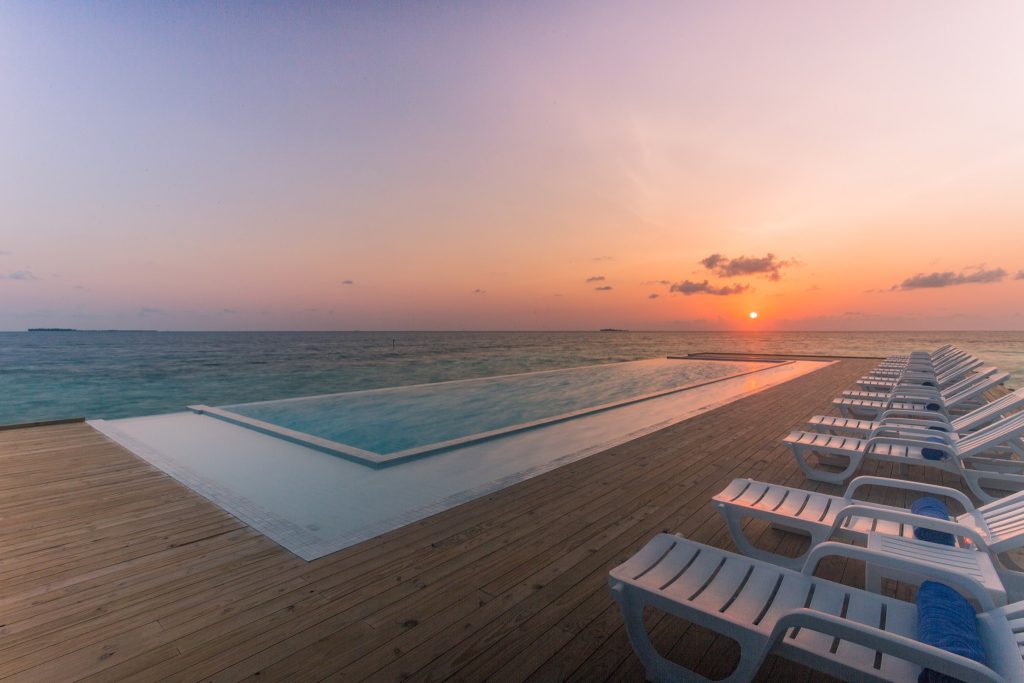 Infinity-Pool mit weißen Liegestühlen und blauen Handtüchern auf Holzterrasse bei Sonnenuntergang auf den