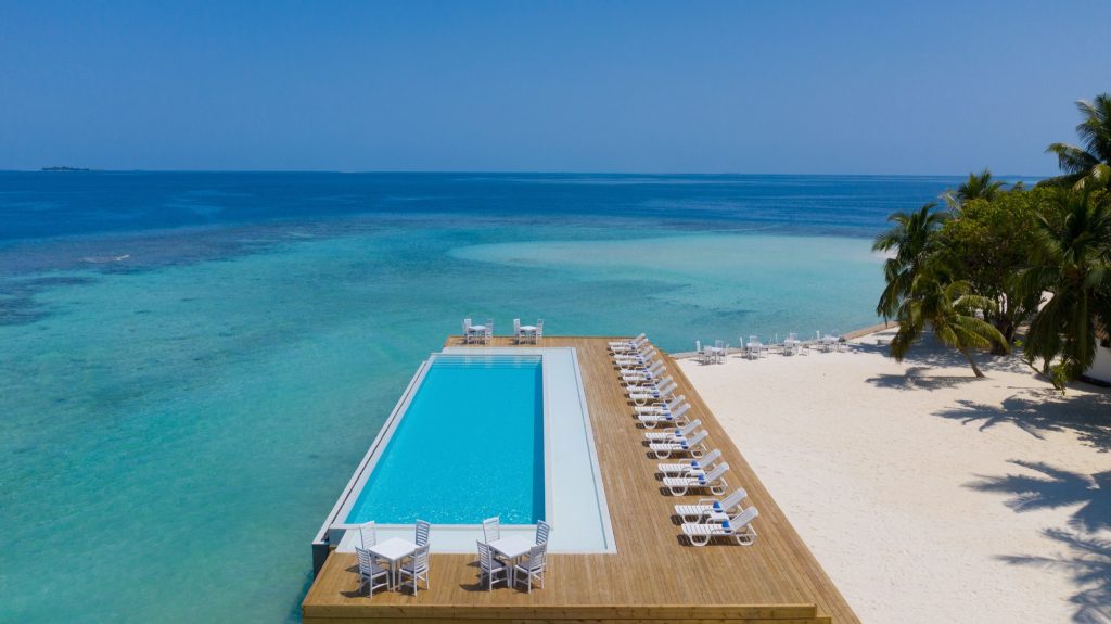 Großer rechteckiger Infinity-Pool mit weißen Liegestühlen und Holzdeck am Sandstrand auf den