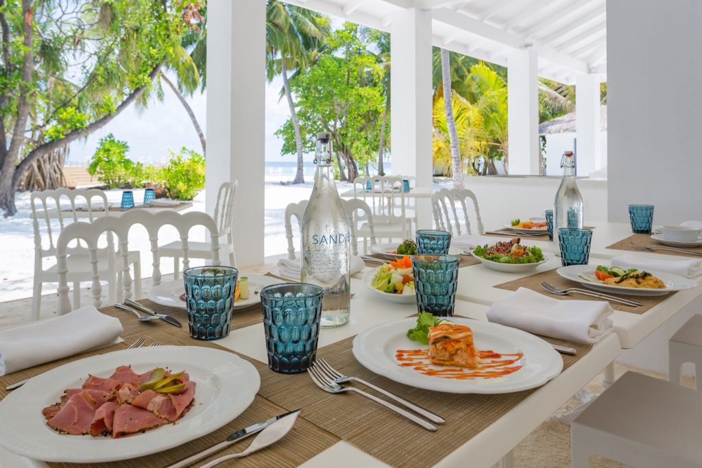Gedeckte Terrasse im Restaurant von Sandies Bathala Maldives mit weißen Tischen, blauen Gläsern und Palmen im Hintergrund