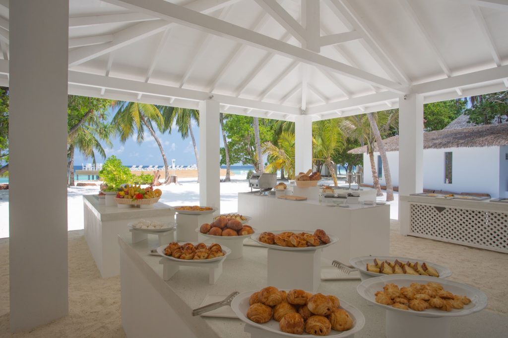 Offener weißer Pavillon im Restaurant Sandies Bathala Maldives mit Tischen, Gebäck, Obst und Blick auf Meer und Palmen