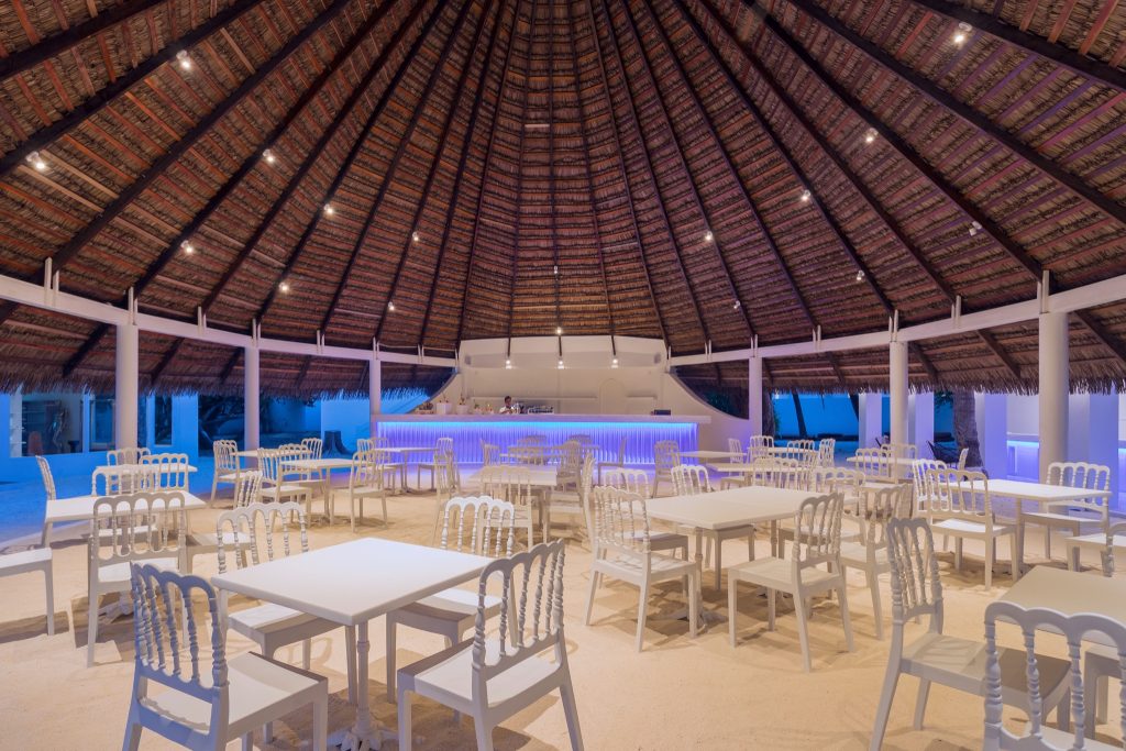 Offene Strandbar mit weißer Einrichtung, Palmdach aus Holz und Stroh, blaue Beleuchtung, Dämmerung Malediven