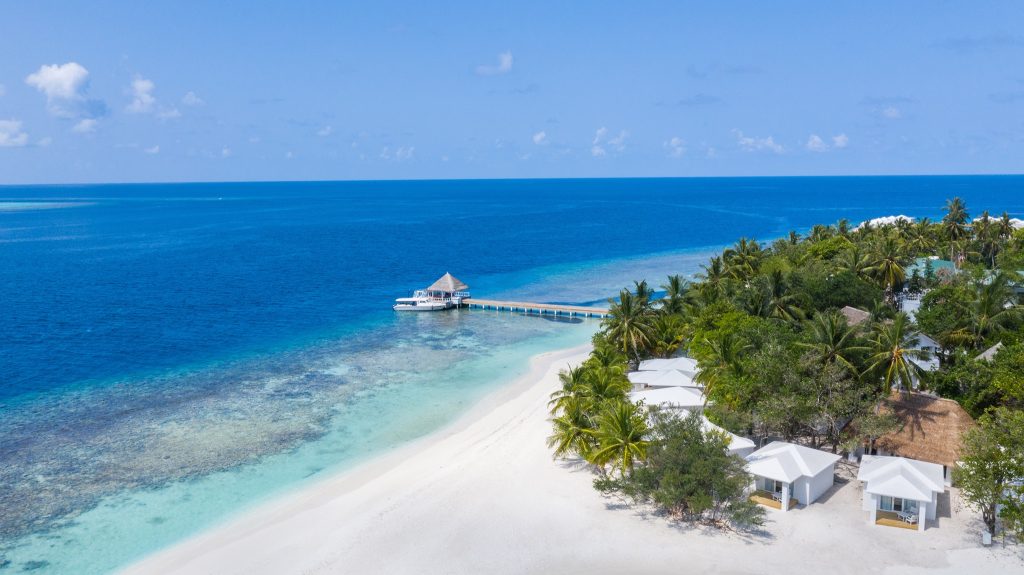 Luftaufnahme einer Resort mit weißen Bungalows am Sandstrand und Holzsteg über türkisblauem Wasser auf den