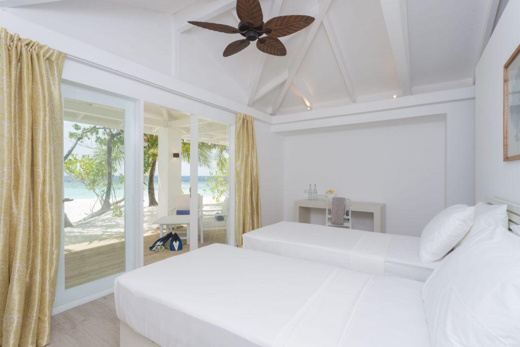 Hotelzimmer mit zwei weißen Einzelbetten, Holzdeckenventilator, gelben Vorhängen und Meerblick auf den
