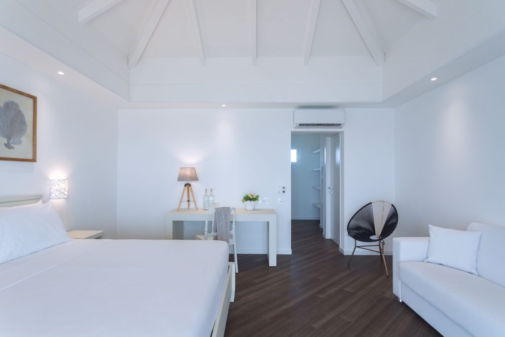 Hotelzimmer mit großem Doppelbett, weißer Bettwäsche, Sofa, schwarzem Stuhl und Holztisch mit Lampen auf den