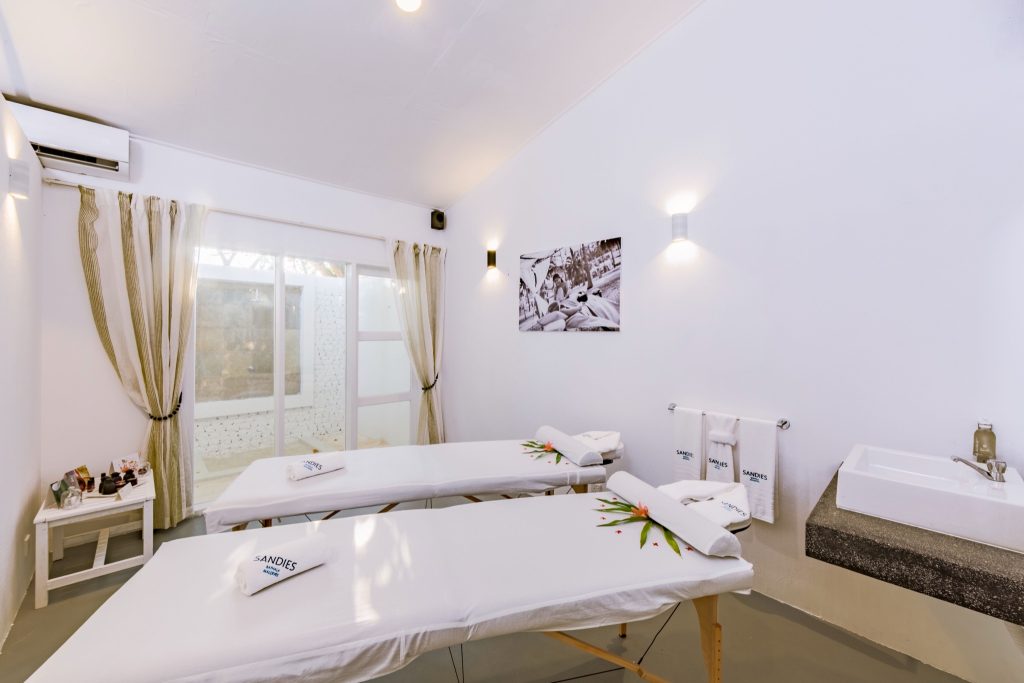 Wellnessraum mit zwei weißen Massageliegen, Steinwaschbecken, Handtüchern und Schwarz-Weiß-Foto an der Wand