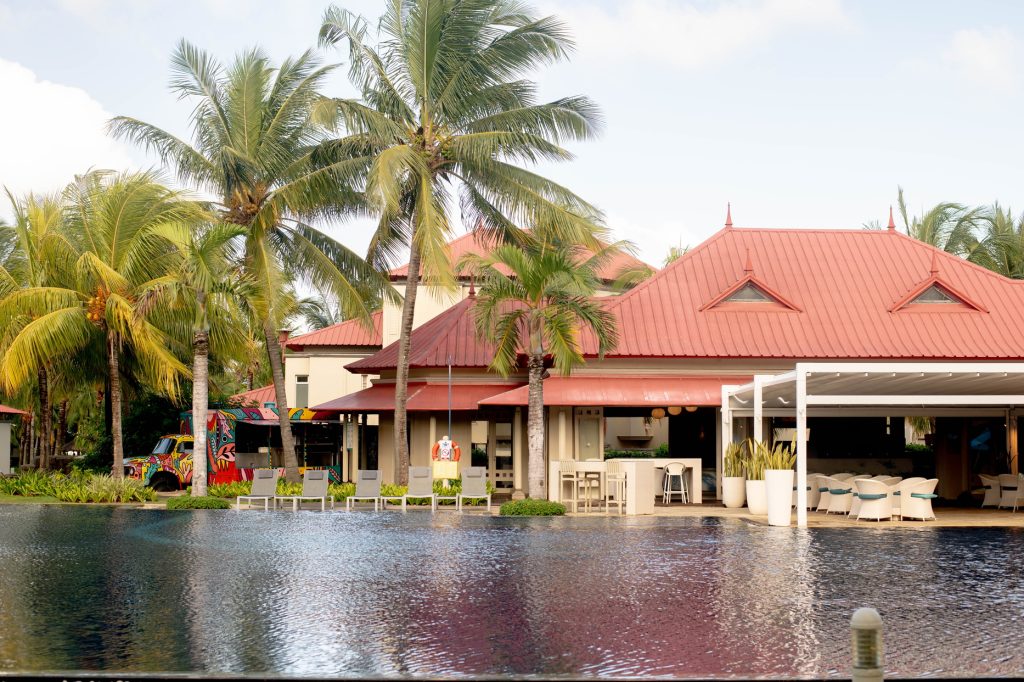 Hotelpool mit roten Dächern, beigen Mauern und Palmen im Tamassa Bel Ombre auf Mauritius