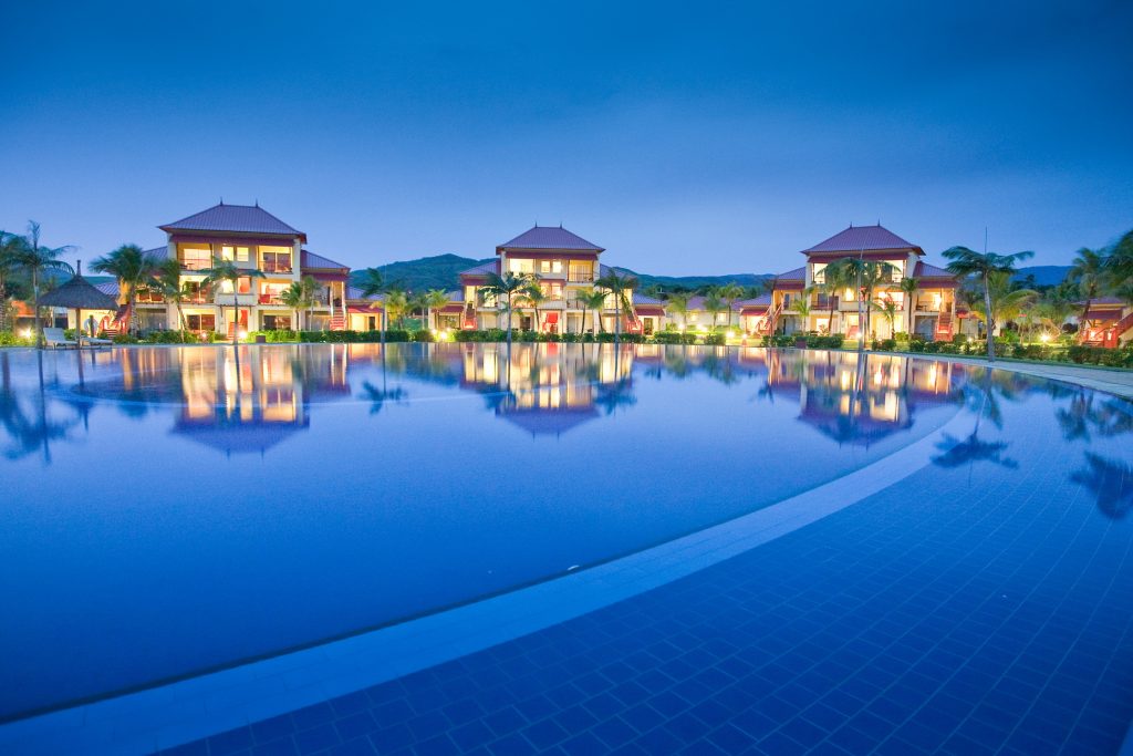 Blick auf Pool mit reflektierendem Wasser, umgeben von zweistöckigen Villen mit roten Dächern und Palmen in Tamassa Bel.