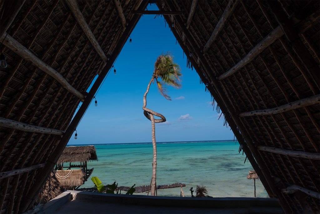 Blick aus offenem dreieckigem Dachfenster einer Holzhütte auf türkisfarbenes Meer und gewundene Palme in Sansibar