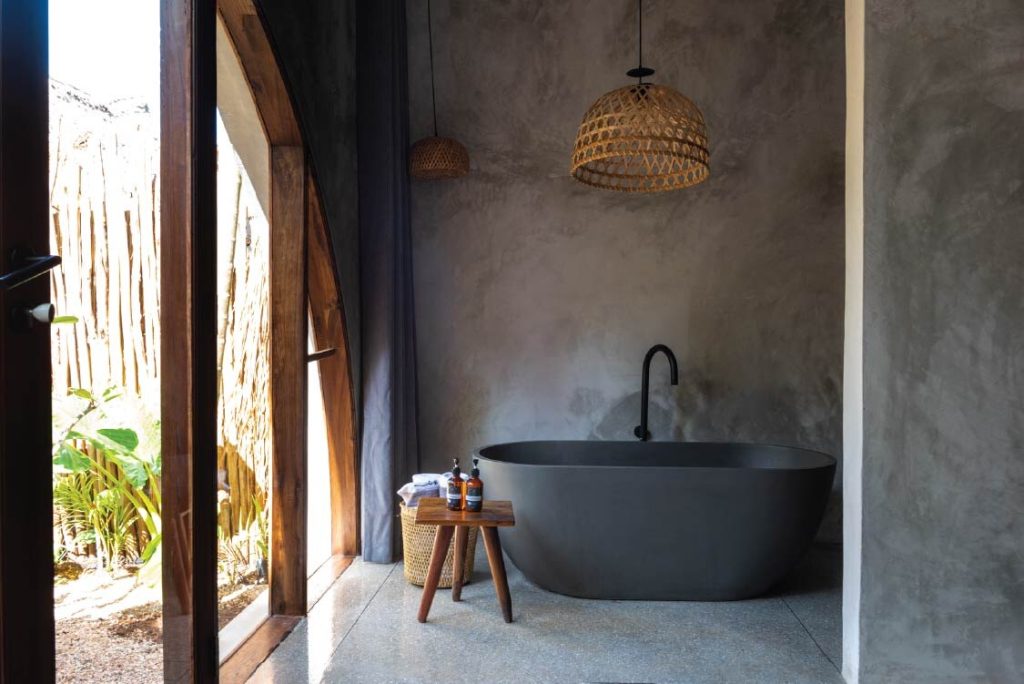 Modernes Badezimmer mit dunkler freistehender Badewanne, Holzhocker, Handtüchern und Glastür zum Außenbereich im Be Zanzibar
