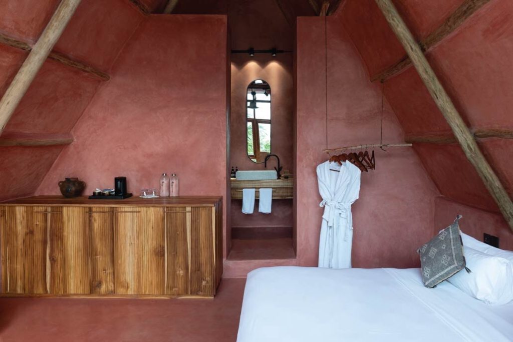 Zimmer im Be Zanzibar Zoe Room mit terrakottafarbenen Wänden, Holzschrank, Bademantel und Badezimmer mit ovalem Spiegel