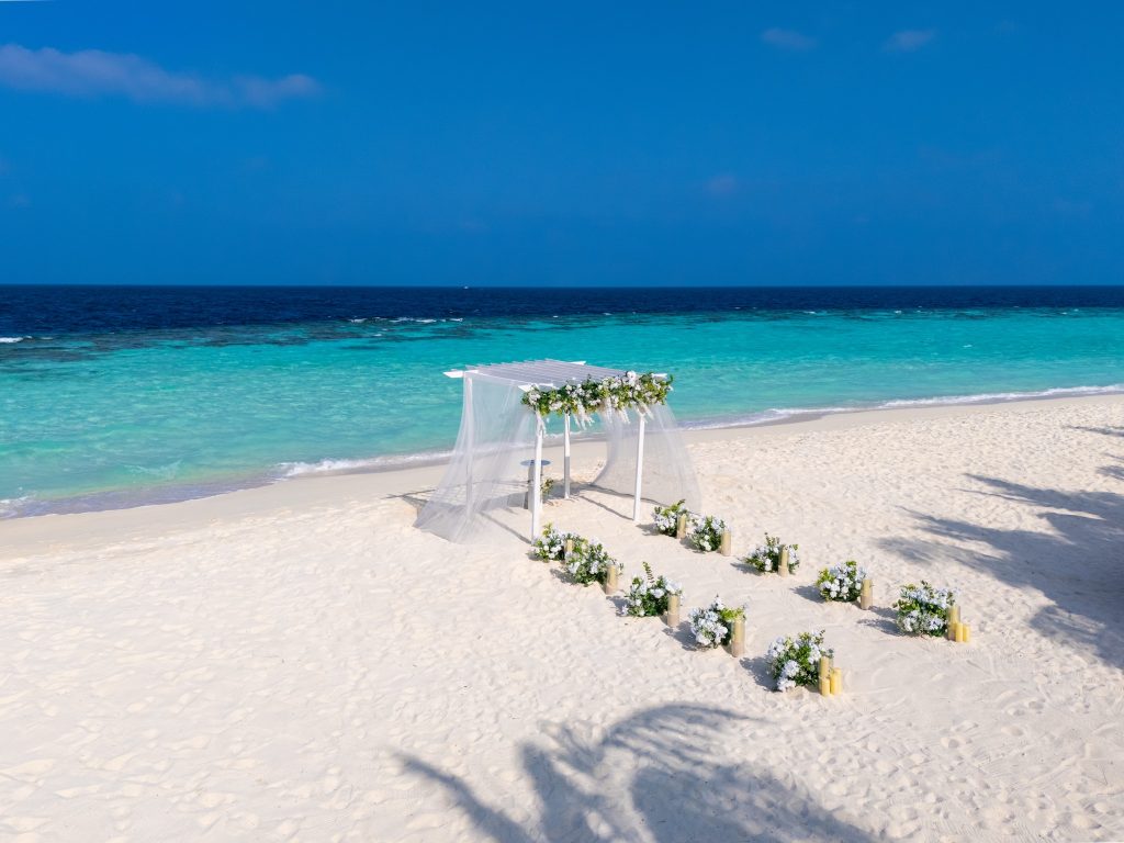 Strandabschnitt mit hellem Sand, hölzernem Pavillon mit weißem Stoffdach und Blumenarrangements auf den