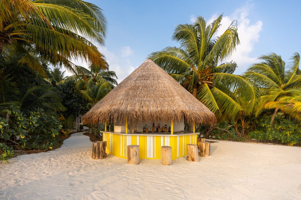 Strandbar mit rundem strohgedecktem Dach, gelb-weiß gestrichenem Holz und groben Holzstämmen als Sitzgelegenheiten.
